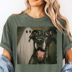 May include: Olive green t-shirt featuring a distressed image of a surprised dog and a ghost. The dog has wide eyes and an open mouth. The ghost is white with two black eye holes. The shirt has short sleeves and a relaxed fit.