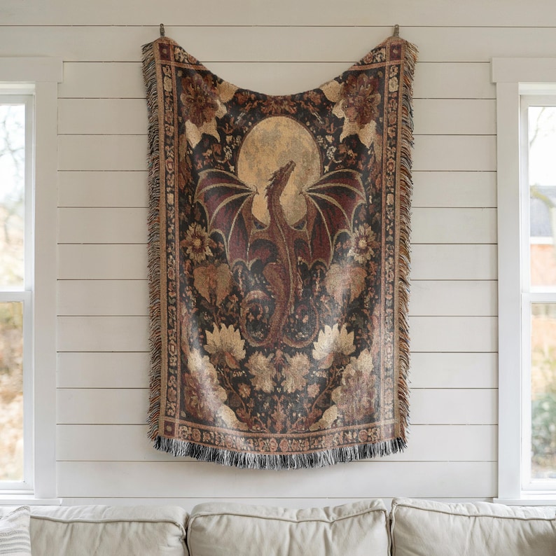 May include: A tapestry-style throw blanket featuring a dragon with outstretched wings against a full moon. The dragon is brown and red, with a black background and floral accents. The blanket has a fringed edge and is hanging on a white wall.
