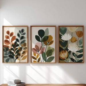 Peut inclure: Trois impressions d'art botanique encadrées. Chaque impression présente des motifs floraux et végétaux stylisés dans des tons de vert, d'orange, de jaune et de rose sur un fond neutre. Les cadres sont en bois. L'œuvre est exposée sur un mur blanc.