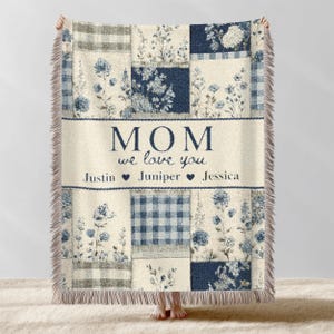 May include: A cream-colored throw blanket with a patchwork design featuring floral and plaid patterns in shades of blue and gray. The blanket has a cream-colored banner with the text "MOM we love you" and names below. The blanket has a fringed edge.
