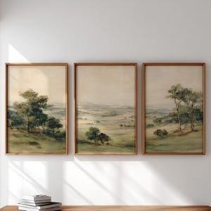 May include: Three framed landscape prints. Each print depicts a watercolour painting of a tranquil, rolling green landscape with trees and a soft, hazy sky. The frames are made of wood.