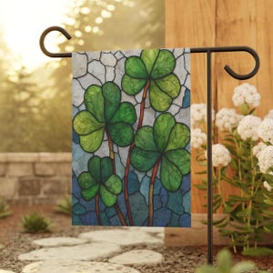 May include: A garden flag featuring a stained-glass design of four green shamrocks with brown stems against a blue and gray background. The flag is displayed on a black metal stand in an outdoor setting.