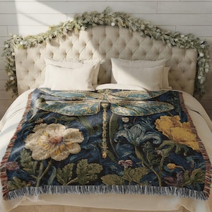 Puede incluir: Una manta decorativa con un dise&ntilde;o de lib&eacute;lula, con un fondo azul oscuro y elementos florales y bot&aacute;nicos en tonos de amarillo, verde y crema. La manta tiene un borde con flecos y se muestra en una cama.