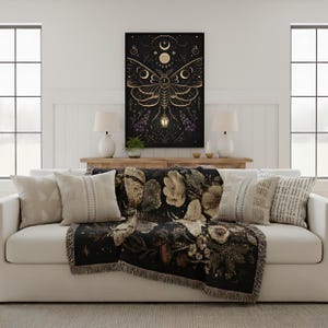May include: A living room scene featuring a white sofa adorned with decorative pillows and a black throw blanket with a floral design. A framed art piece with a moth illustration hangs above the sofa. Two white lamps sit on a wooden console.