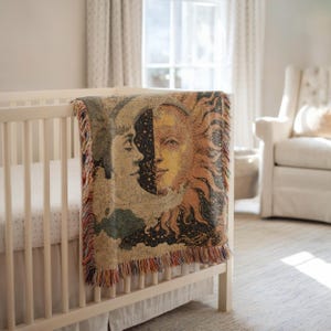 May include: A woven tapestry blanket draped over a white crib. The blanket features a celestial design with a sun and moon motif in shades of gold, orange, and blue. The blanket has a fringed edge and is made of a soft, textured material.