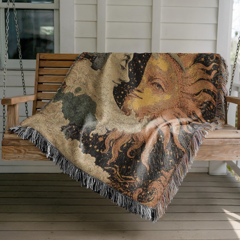 May include: Woven throw blanket featuring a celestial design with a sun and moon motif. The blanket has a fringed edge and is draped over a wooden swing. The color palette includes shades of tan, brown, black, and gold.