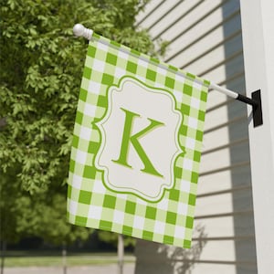 May include: A green and white checkered garden flag with a large, light green letter "K" inside a decorative frame. The flag is hanging on a white pole attached to a white wall. The flag is made of fabric and is designed for outdoor use.