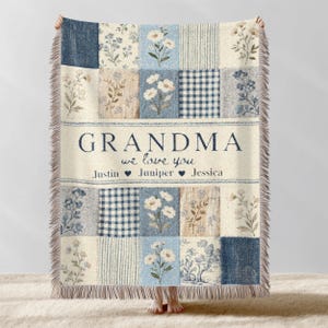 以下が含まれることがあります： クリーム色のフリンジが付いたパッチワークのスローブランケット。ブランケットは、さまざまな青とクリーム色の花柄とギンガムチェック柄が特徴です。「GRANDMA we love you」というテキストが中央に表示され、その下に名前が表示されています。