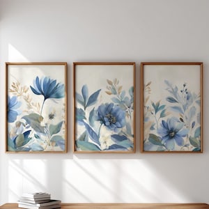 May include: Three framed floral art prints. Each print features watercolor-style blue flowers, green leaves, and beige accents against a cream background. The frames are made of light brown wood. The artwork is displayed on a white wall.