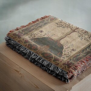 Peut inclure: Une couverture pli&eacute;e en tapisserie avec un motif d'arbre dans les tons marron, vert et bordeaux. La couverture a une bordure &agrave; franges noire, rouge et marron. Le plaid est pli&eacute; sur une surface en bois.