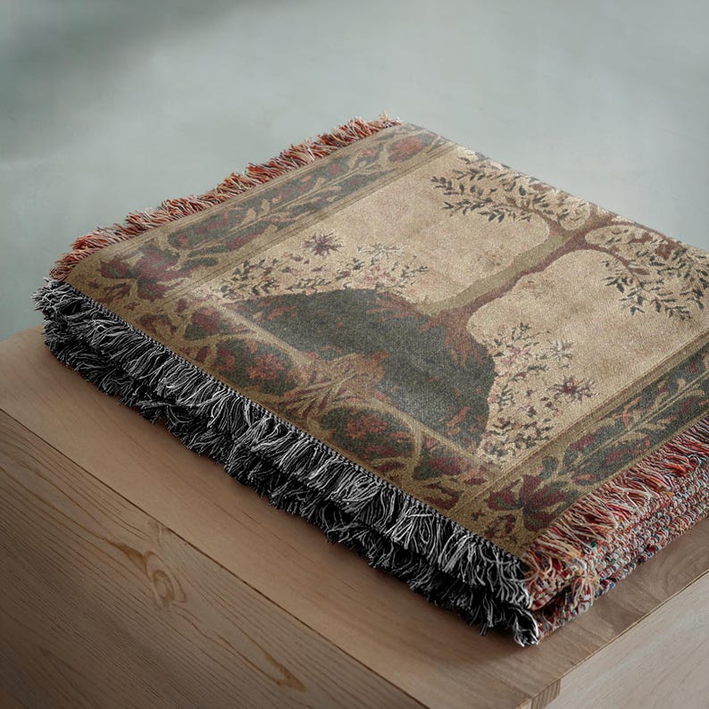 Peut inclure: Une couverture pli&eacute;e en tapisserie avec un motif d'arbre dans les tons marron, vert et bordeaux. La couverture a une bordure &agrave; franges noire, rouge et marron. Le plaid est pli&eacute; sur une surface en bois.