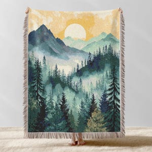 May include: A woven throw blanket featuring a watercolor landscape of a forest and mountains under a yellow sun. The blanket has a fringe border and is primarily green, blue, and yellow.