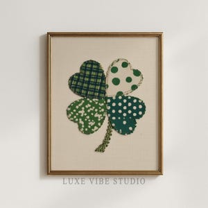 May include: Framed artwork featuring a four-leaf clover design. The clover's four heart-shaped leaves are in green patterns: plaid, polka dots, and floral. The artwork is set against a cream background and framed in gold. The text "LUXE VIBE STUDIO" is at the bottom.