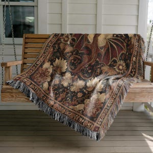 May include: A woven throw blanket with a dragon and floral design in shades of brown, black, and tan. The blanket is draped over a wooden porch swing and has a fringed edge.