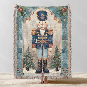 Puede incluir: Una manta tejida con una ilustración detallada de un soldado cascanueces con uniforme azul. El diseño incluye árboles de Navidad, copos de nieve y bordes decorativos en tonos azules, beige y rojos. La manta tiene un borde con flecos.