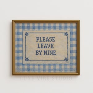 May include: A framed artwork with a gold-coloured frame. The image shows a blue and white gingham pattern background. The centre has a cream-coloured rectangle with the text "PLEASE LEAVE BY NINE" in blue cross-stitch style.