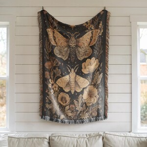 May include: A dark gray throw blanket with a botanical and insect design. The blanket features two large moths and floral elements in shades of tan and brown. The blanket has a fringed edge and is hanging on a white wall.