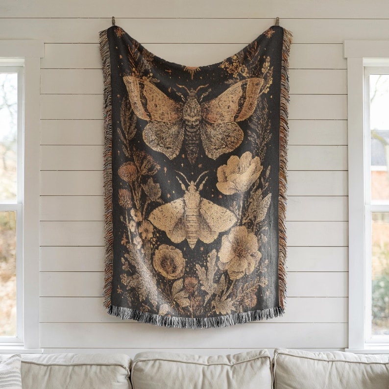 May include: A dark gray throw blanket with a botanical and insect design. The blanket features two large moths and floral elements in shades of tan and brown. The blanket has a fringed edge and is hanging on a white wall.