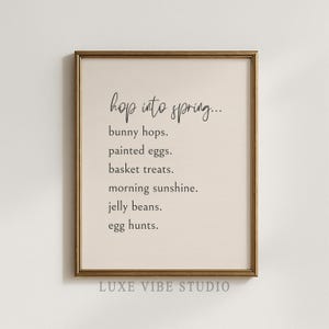 May include: Framed print with the text "hop into spring..." followed by a list of spring-themed items: bunny hops, painted eggs, basket treats, morning sunshine, jelly beans, and egg hunts. The print is in a gold frame.