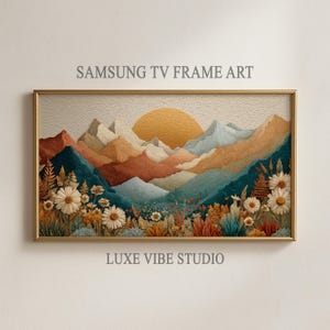 May include: A framed landscape art print for a Samsung TV. The artwork depicts a mountain range with a setting sun, in shades of orange, teal, and beige. White daisies and other plants are in the foreground. Text reads "SAMSUNG TV FRAME ART" and "LUXE VIBE STUDIO."