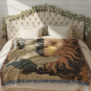 May include: A woven throw blanket featuring a celestial design with a sun and moon motif. The blanket displays a detailed illustration in shades of beige, brown, and black, with a fringed edge. The blanket is placed on a bed with white linens.