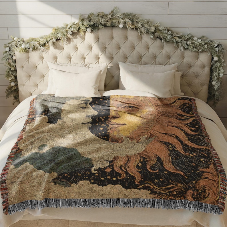 May include: A woven throw blanket featuring a celestial design with a sun and moon motif. The blanket displays a detailed illustration in shades of beige, brown, and black, with a fringed edge. The blanket is placed on a bed with white linens.