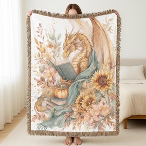 Könnte beinhalten: Eine gewebte Decke mit einer Aquarellillustration eines goldenen Drachen, der ein Buch liest. Der Drache ist von Sonnenblumen, Kürbissen und Herbstlaub in Gold-, Orange- und Grüntönen umgeben. Die Decke hat einen Fransenrand.