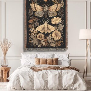 May include: A black tapestry featuring two moths and floral elements in shades of gold and beige. The tapestry is rectangular with a fringed edge, hanging above a bed with white bedding and brown pillows. The overall theme is nature and bohemian decor.