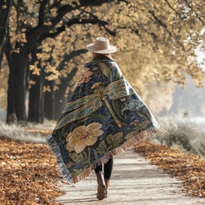 Puede incluir: Una persona con sombrero beige y una manta azul oscuro con un gran dise&ntilde;o de lib&eacute;lula y flores. La manta tiene un borde con flecos y est&aacute; drapeada sobre los hombros de la persona que camina por un camino bordeado de &aacute;rboles.
