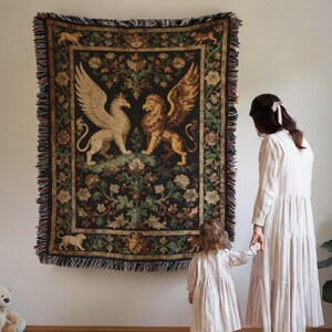 May include: A tapestry featuring a griffin and a lion with large wings, set against a dark background with floral and foliage patterns. The tapestry has a fringed border and is displayed on a white wall.
