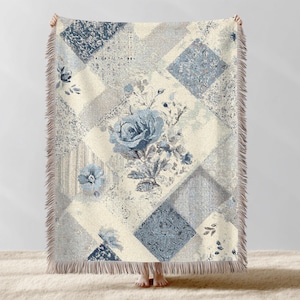 Puede incluir: Una manta con diseño de patchwork en tonos azules, grises y crema. La manta presenta patrones florales, incluyendo una gran rosa azul, y formas geométricas. Los bordes están acabados con flecos.