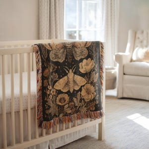 May include: A dark gray blanket with a moth and floral design draped over a white crib. The blanket features a large moth in the center, surrounded by flowers and foliage in shades of tan and cream. The blanket has a fringed edge with red, orange, and brown threads.