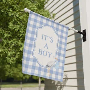 May include: A blue and white gingham pattern flag with the text "IT'S A BOY" in a white decorative frame. A stork carrying a blue bundle is featured below the text. The flag is mounted on a white pole.