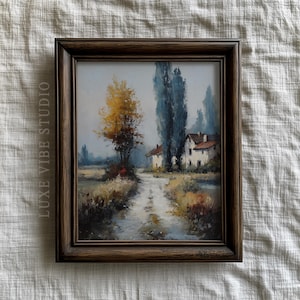 May include: Framed landscape painting featuring a dirt road leading to white houses with red roofs. The artwork depicts trees with yellow and blue foliage, set against a muted sky. The frame is dark brown, and the text "LUXE VIBE STUDIO" is visible on the left side.