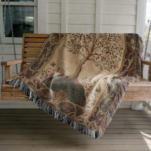 Peut inclure: Une couverture beige avec un motif d'arbre drap&eacute;e sur une balan&ccedil;oire en bois. L'arbre a des branches marron et des feuilles vertes. La couverture a une bordure d&eacute;corative avec des franges.