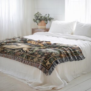 May include: A decorative throw blanket with a detailed design featuring a griffin, floral patterns, and a fringed edge. The blanket is primarily black, with accents of gold, green, and brown. It is draped over a white bed, creating a cozy and elegant aesthetic.
