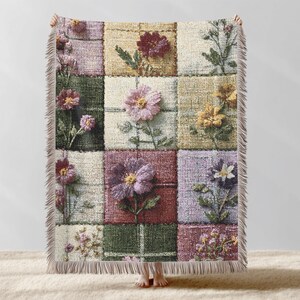 May include: A patchwork throw blanket featuring embroidered flowers in various colors, including pink, purple, and yellow. The blanket has a fringed edge and is composed of squares in shades of green, beige, and burgundy. The floral design adds a touch of nature to the home decor.