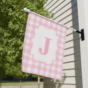 May include: A pink and white gingham flag featuring a white scalloped border and a pink letter "J". The flag is mounted on a white pole with a black bracket. The flag is displayed outside.