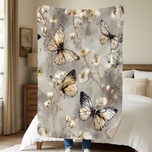 May include: A gray blanket featuring a watercolor design of butterflies and floral elements. The butterflies have tan and black wings, and the flowers are white and gold. The blanket is held up in front of a bed.