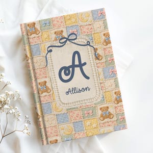 Pode incluir: Um livro de memórias de bebé personalizado com um design de patchwork. A capa apresenta ursinhos de peluche, luas, estrelas e laços em cores pastel. Uma grande letra azul "A" e o nome "Allison" estão centrados.