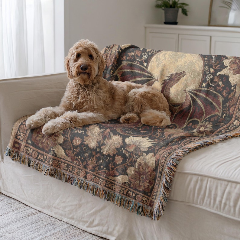 May include: A decorative tapestry throw blanket with a dragon and moon design in shades of black, brown, and gold. The blanket has a fringed edge and is draped over a white sofa. A golden doodle dog is lying on the sofa.