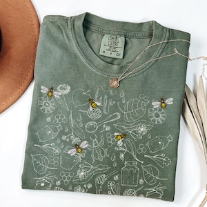 May include: A green t-shirt with a white design of bees, honeycombs, and flowers. The text "COMFORT COLORS" is on the tag.