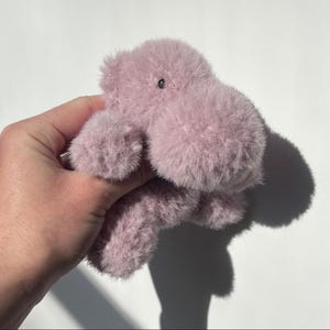 May include: A plush, lavender-colored hippo toy with a soft, furry texture. The toy has a small, black eye and is held against a white background. The hippo's shadow is visible.