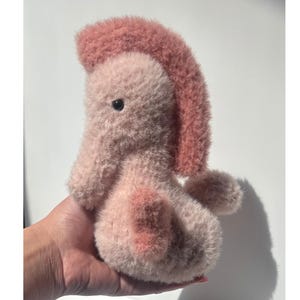 Op de afbeelding: Een handgemaakt pluche wezen in roze en roze tinten. Het zachte speelgoed heeft een getuften kuif en een afgerond lichaam. Het wezen wordt in een hand gehouden tegen een witte achtergrond, met een schaduw naar rechts.
