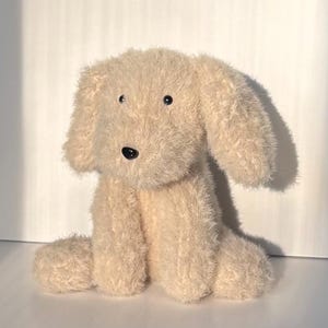 May include: A plush dog toy made of light beige, shaggy, textured fabric. The stuffed animal features two long, floppy ears, a small black plastic nose, and two small black plastic eyes positioned on the front of its rounded head.