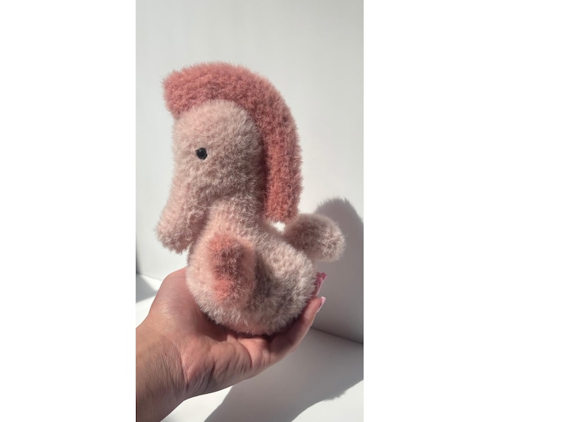 Op de afbeelding: Een handgemaakt pluche zeepaardje in roze tinten. Het zeepaardje heeft een zachte textuur, met een donkerroze manen en accenten op zijn lichaam. Het heeft een klein, donker oog en wordt in een hand gehouden.
