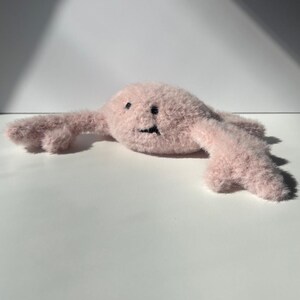 May include: A light pink plush toy with a smiling face and outstretched arms. The toy has a fuzzy texture and black embroidered eyes and mouth. The toy is lying on a white surface.