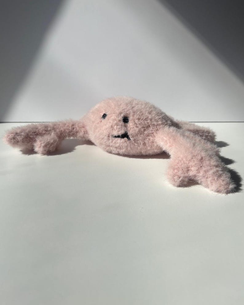 May include: A light pink plush toy with a smiling face and outstretched arms. The toy has a fuzzy texture and black embroidered eyes and mouth. The toy is lying on a white surface.