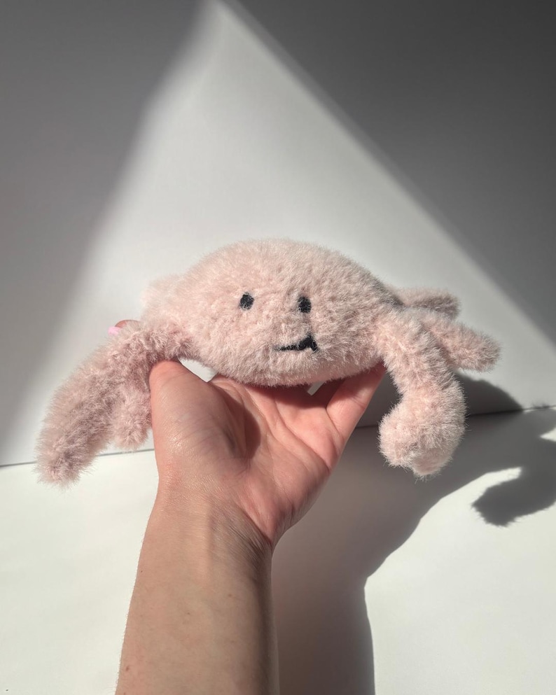 May include: A plush, pink crab toy with a smiling face and embroidered features. The crab has soft, fuzzy fur and is held in a hand against a white background. The toy's claws and body are visible, showcasing its playful design.