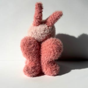 May include: A plush bunny toy in shades of pink. The bunny has long, upright ears and a fluffy body. The fur is soft and textured, with a lighter pink on the face and a darker pink on the ears and body.